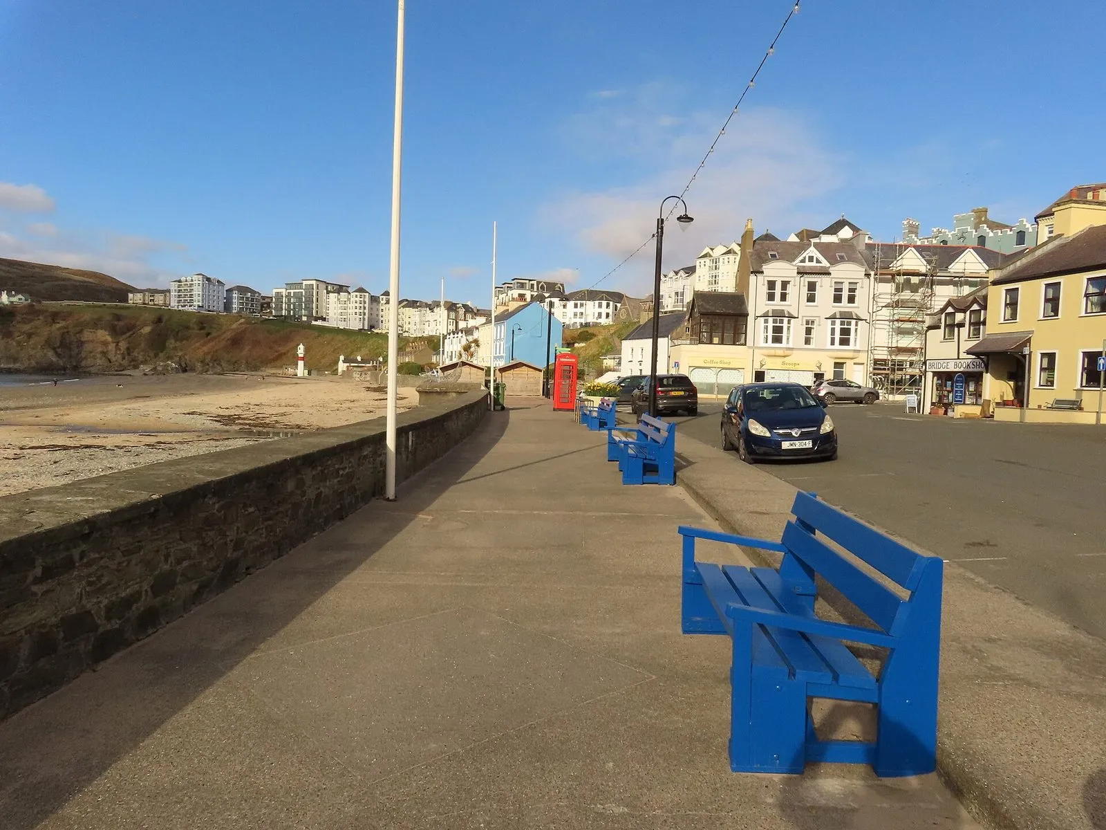 A wide, level concrete promenade in Port Erin with blue benches, a stone wall above the sandy beach and seafront houses behind
