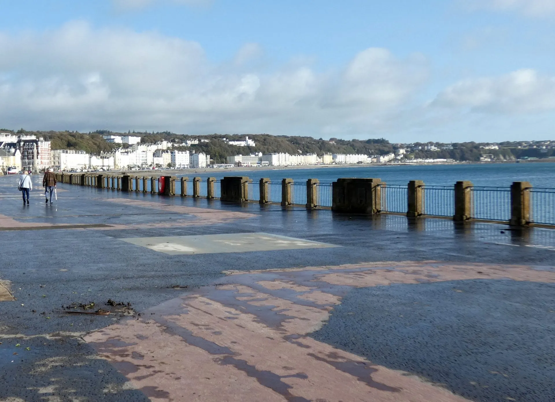 The wide, flat Loch Promenade walkway in Douglas curving along the bay beside iron railings, with white seafront hotels in the distance under a blue sky