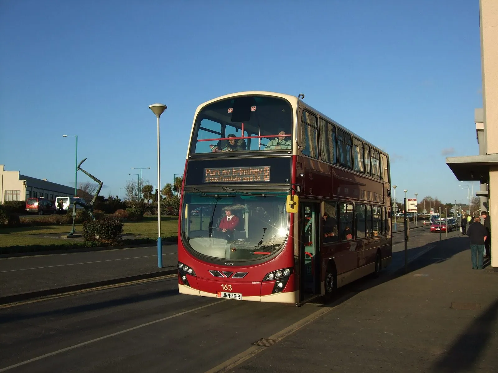 A red Bus Vannin double-decker showing route 8 to Peel waiting at the stand outside Ronaldsway Airport under a clear blue sky