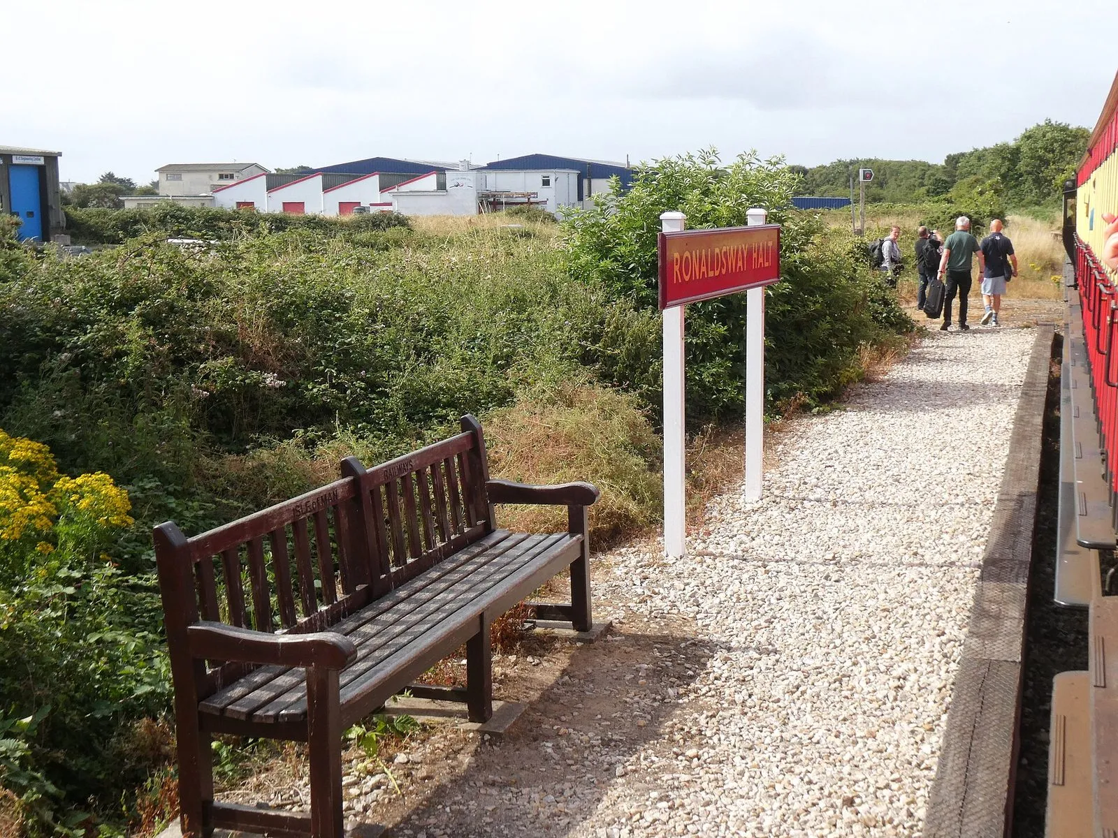 A small gravel platform with a red Ronaldsway Halt sign and a wooden bench, with steam railway carriages alongside and passengers walking away