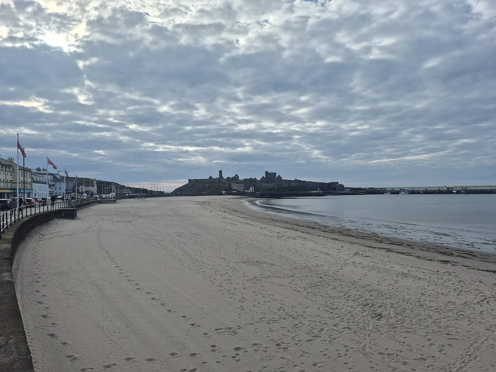 A wide, empty sandy beach curving round Peel Bay towards the castle ruins on the headland, under a cloudy sky