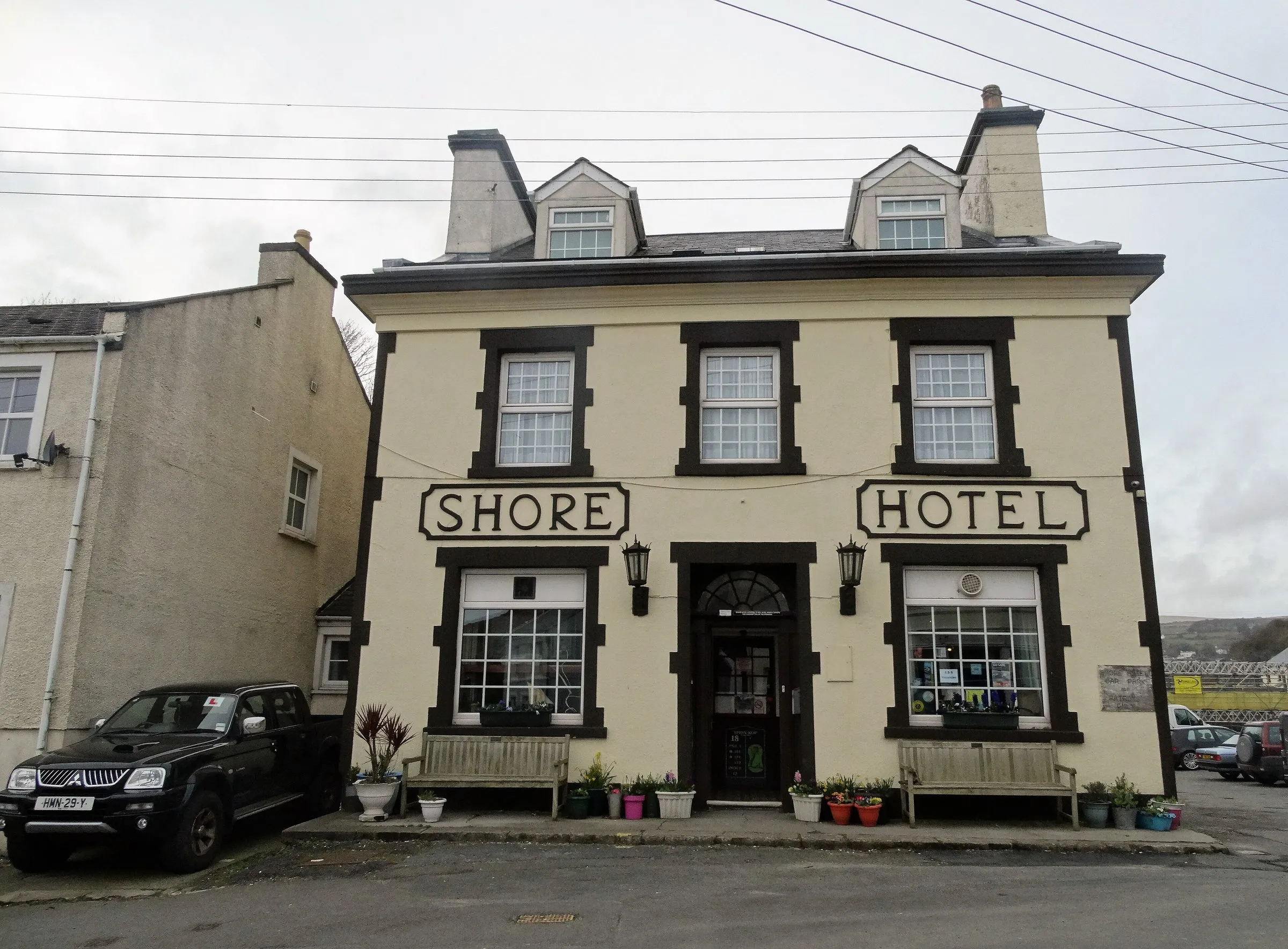 The cream-painted Shore Hotel in Old Laxey, with SHORE HOTEL in black letters above the ground-floor windows and wooden benches out front