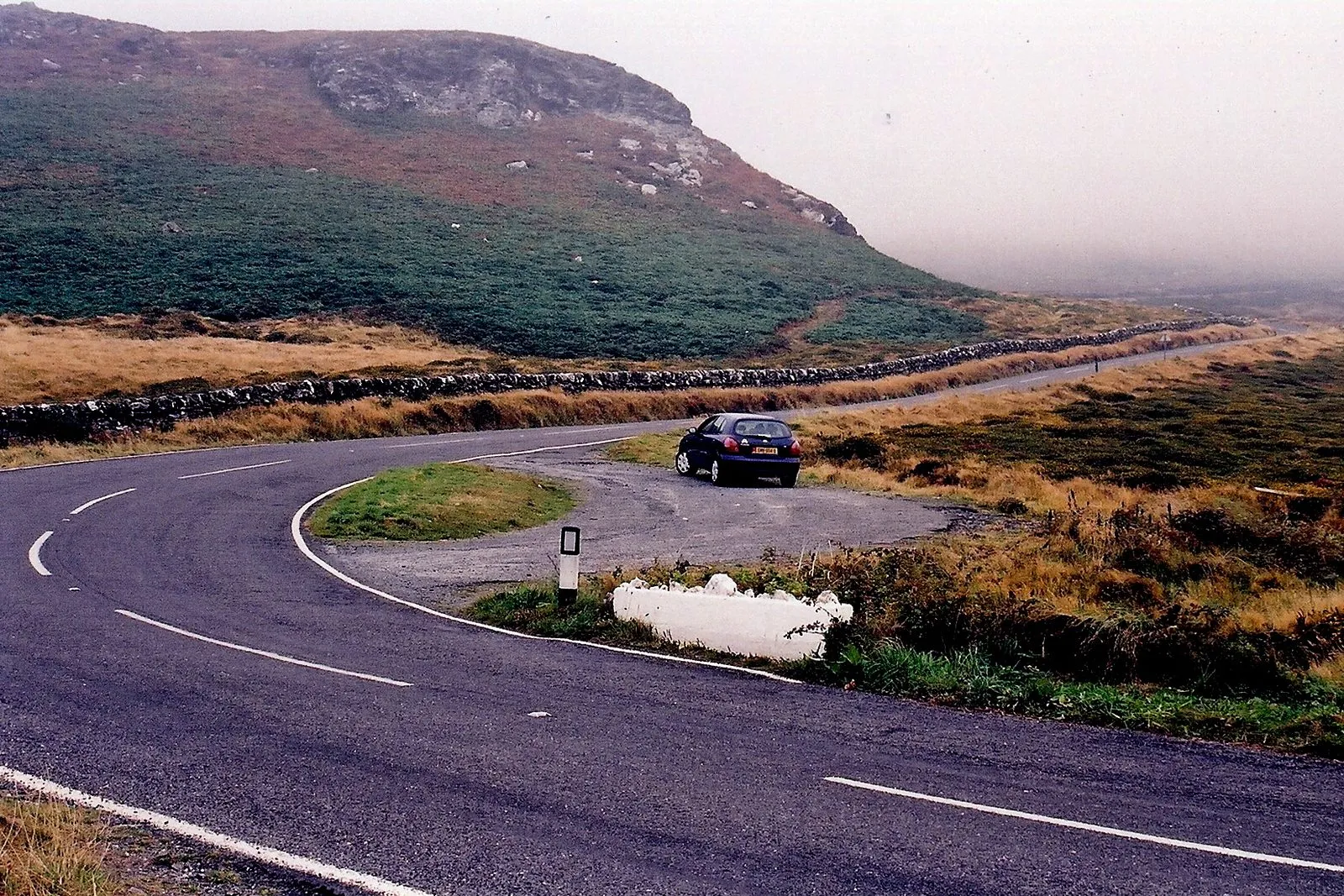 A hairpin bend on the Sloc road with a small lay-by, stone walls and heather on the hillside above, on a misty day
