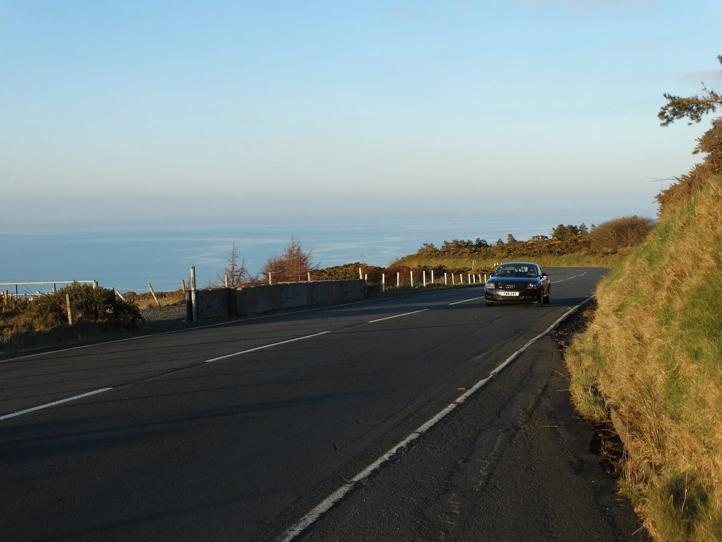 The Mountain Road sweeping round a bend high above the sea near the Gooseneck, with a single car on an otherwise empty road in low evening sun