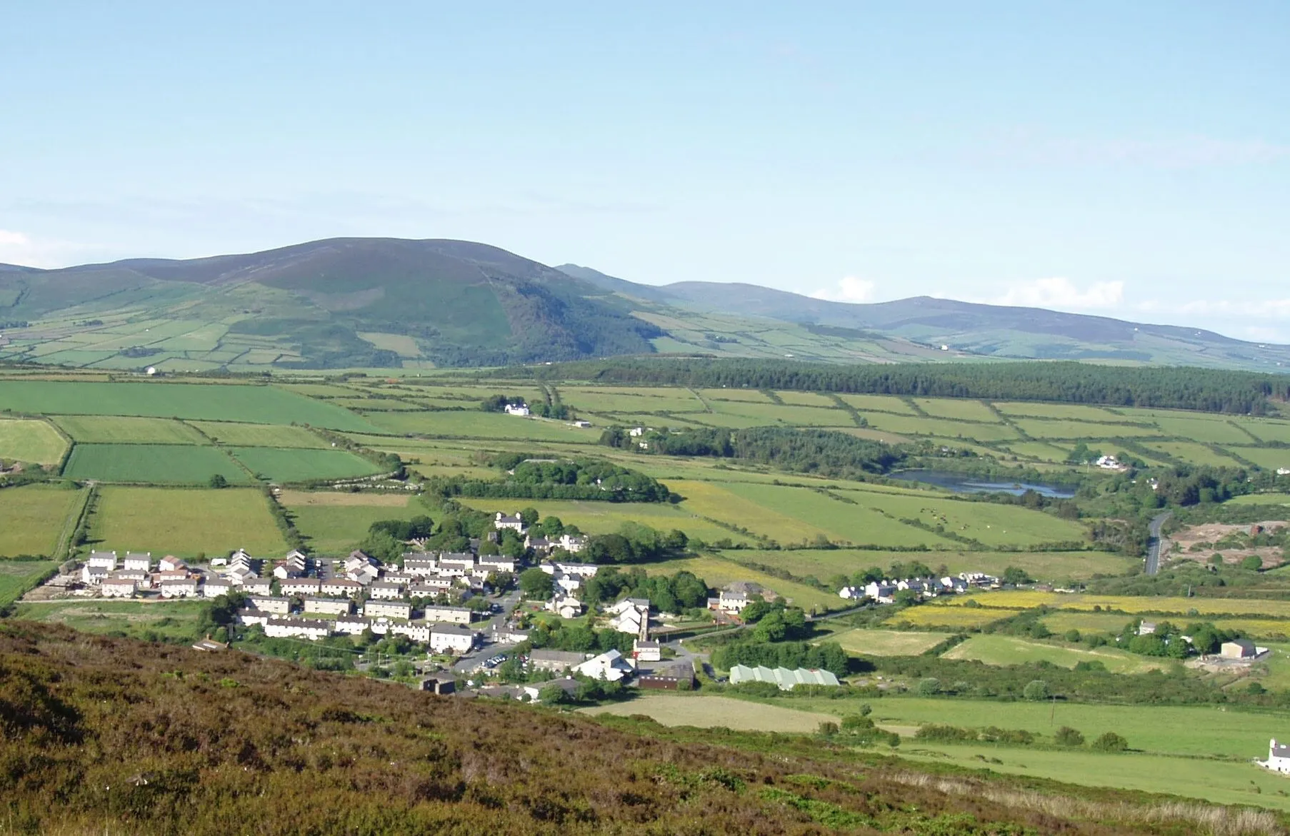 Foxdale village among green fields in the south-central Isle of Man, with hills and a small reservoir behind