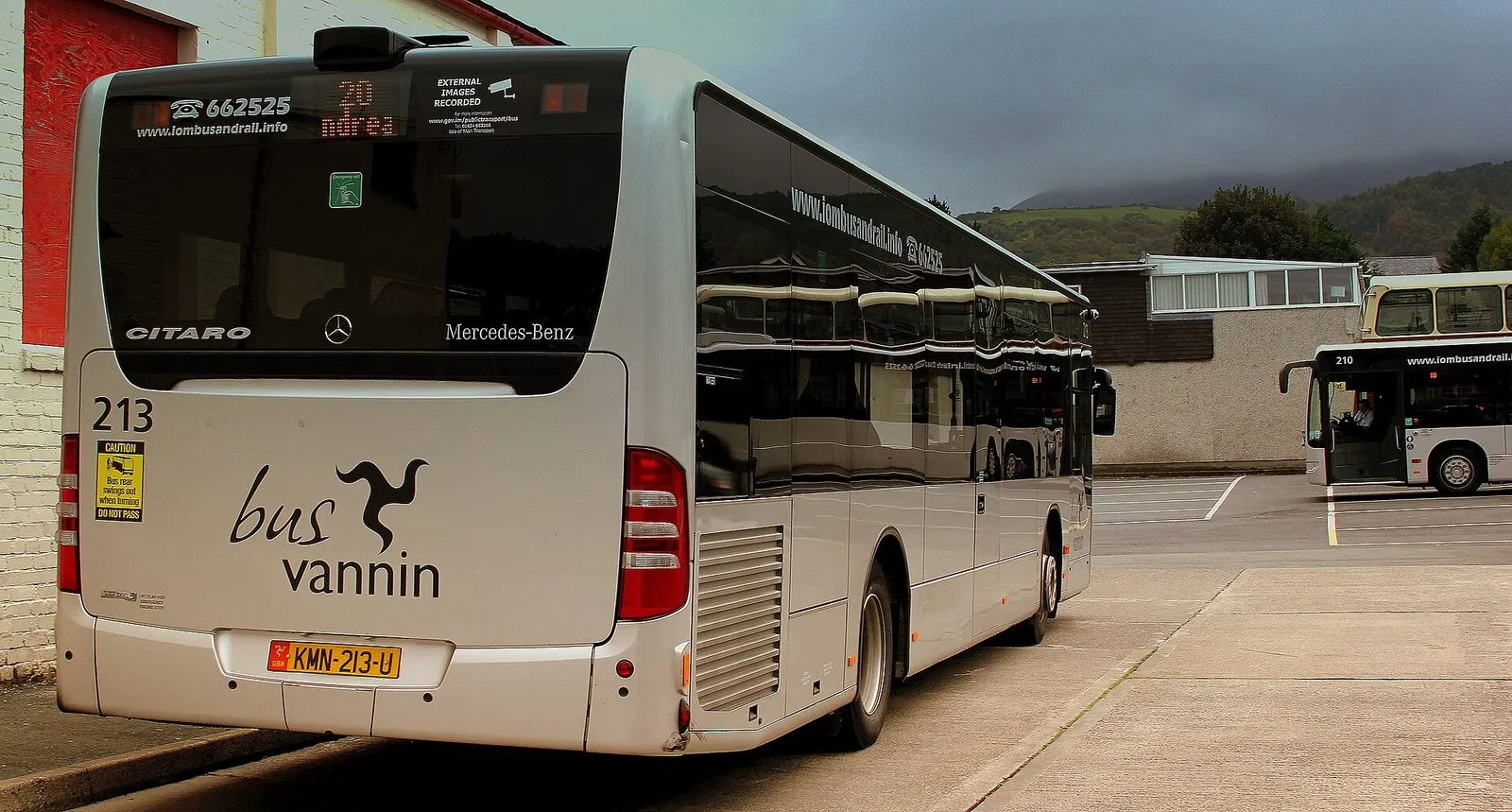 A silver Bus Vannin single-decker bus, route 20 to Andreas, at Ramsey bus station with green hills in low cloud behind
