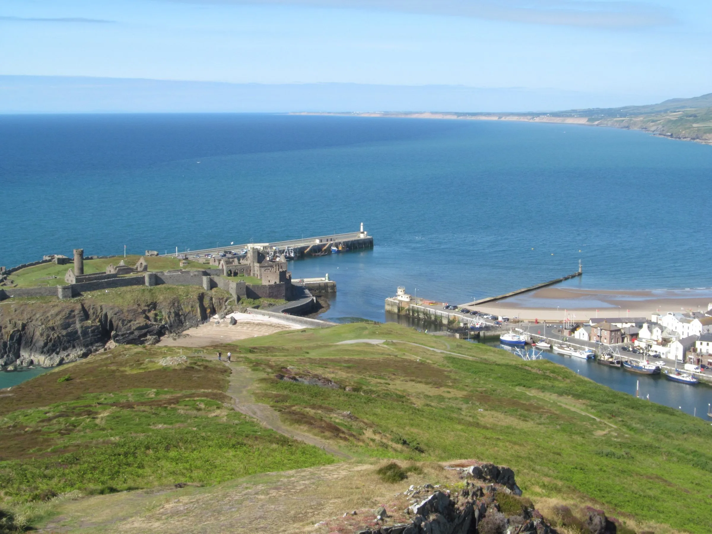 Peel from the slopes of Peel Hill on a sunny day, with the ruins of Peel Castle on St Patrick's Isle, the harbour, the breakwater and the sandy beach curving away up the west coast