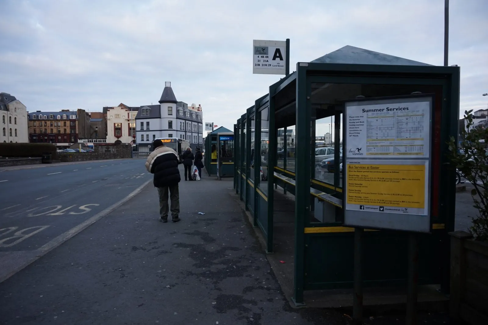 A row of green bus shelters on Lord Street in Douglas, with a stand A sign and a summer timetable poster, and people waiting