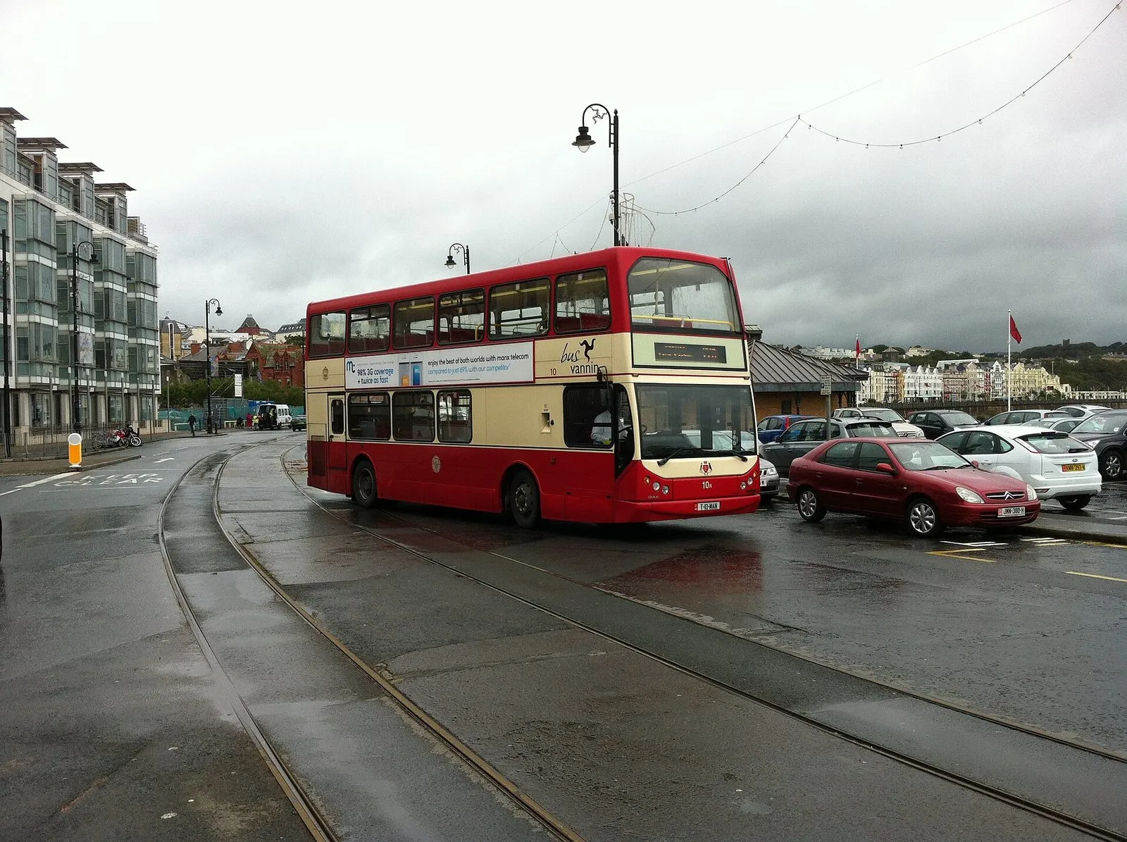 A red and cream Bus Vannin double-decker on the wet promenade at Douglas, crossing the horse tram tracks, with parked cars and flags behind