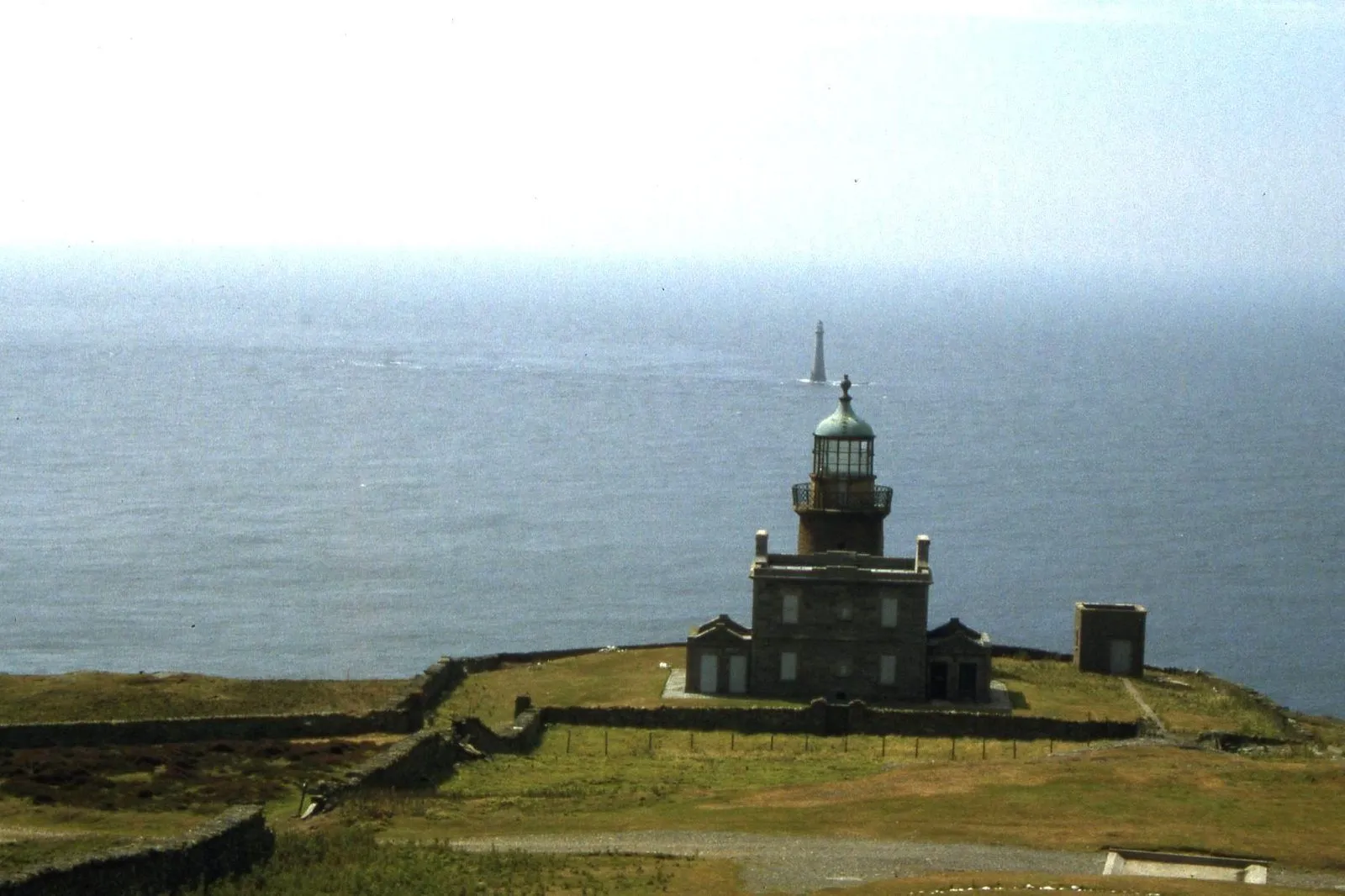 A stone lighthouse with a green-domed lantern on the grassy southern end of the Calf of Man, with Chicken Rock’s tall tower out at sea behind