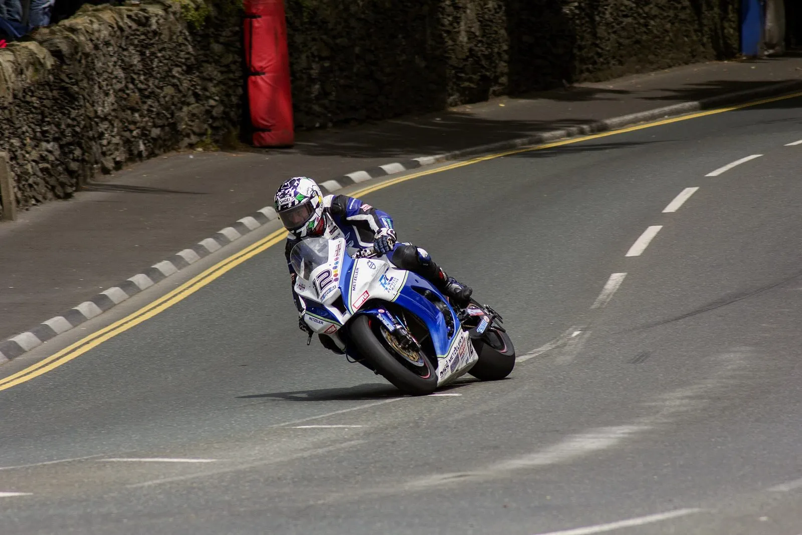 A TT rider leaning a blue and white superbike through the Quarterbridge corner in Douglas, with stone walls and air fencing behind