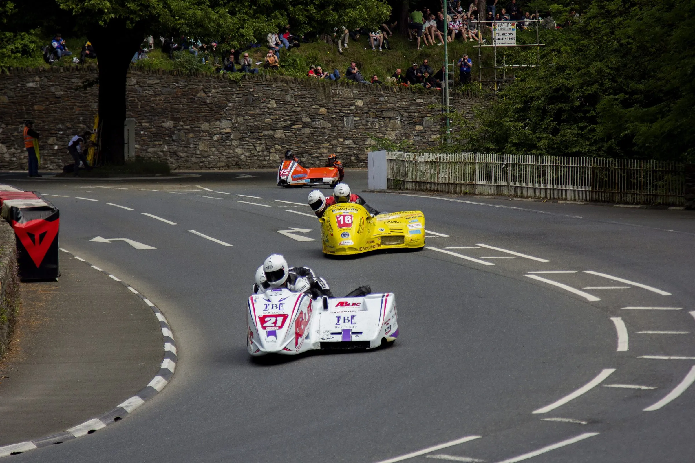 Three racing sidecars rounding the bend at Braddan Bridge, with spectators sitting on the grassy bank above a stone wall