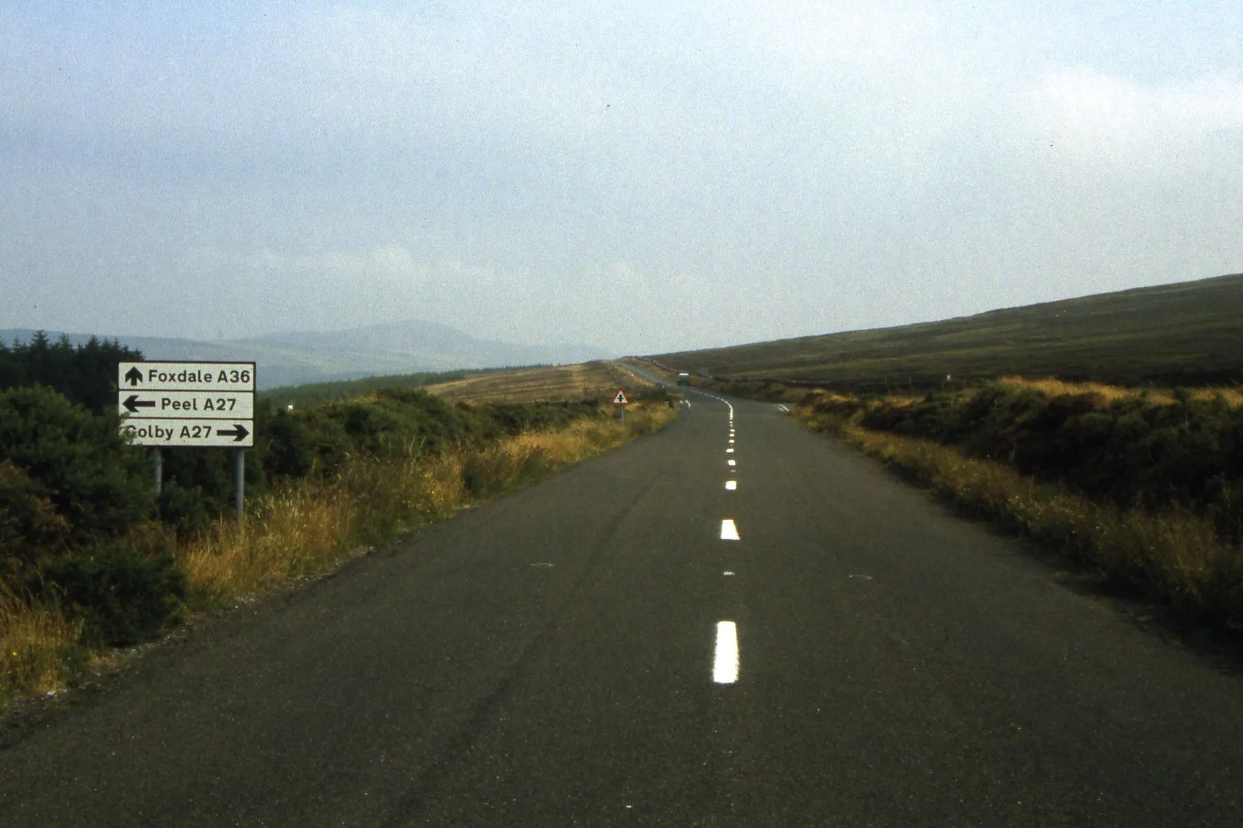 An empty upland road running across moorland towards the Round Table crossroads, with a signpost for Foxdale, Peel and Colby