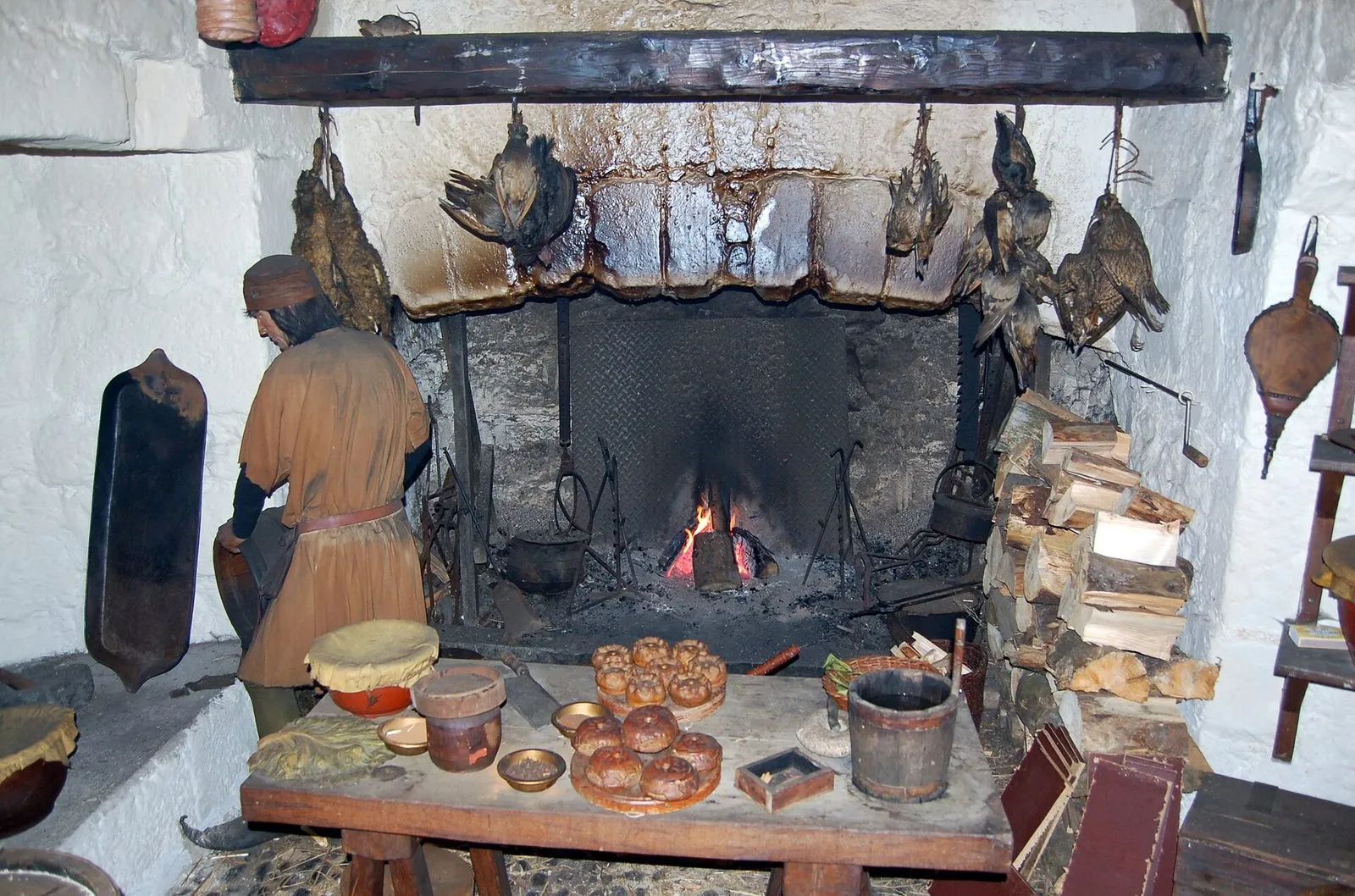A costumed figure by the open fire in Castle Rushen’s medieval kitchen, with game birds hanging from the beam and a table of pies in front