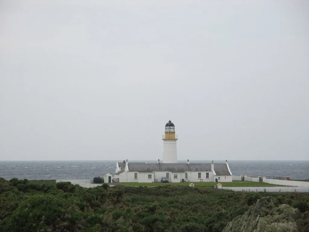 Langness lighthouse, a white tower with a black lantern, behind a row of low white cottages and gorse, with a grey sea beyond