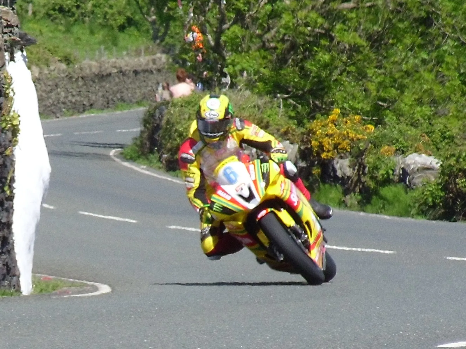 A racer in yellow leaning through a left-hand bend at Tower Bends, between a stone wall and gorse, on the climb out of Ramsey