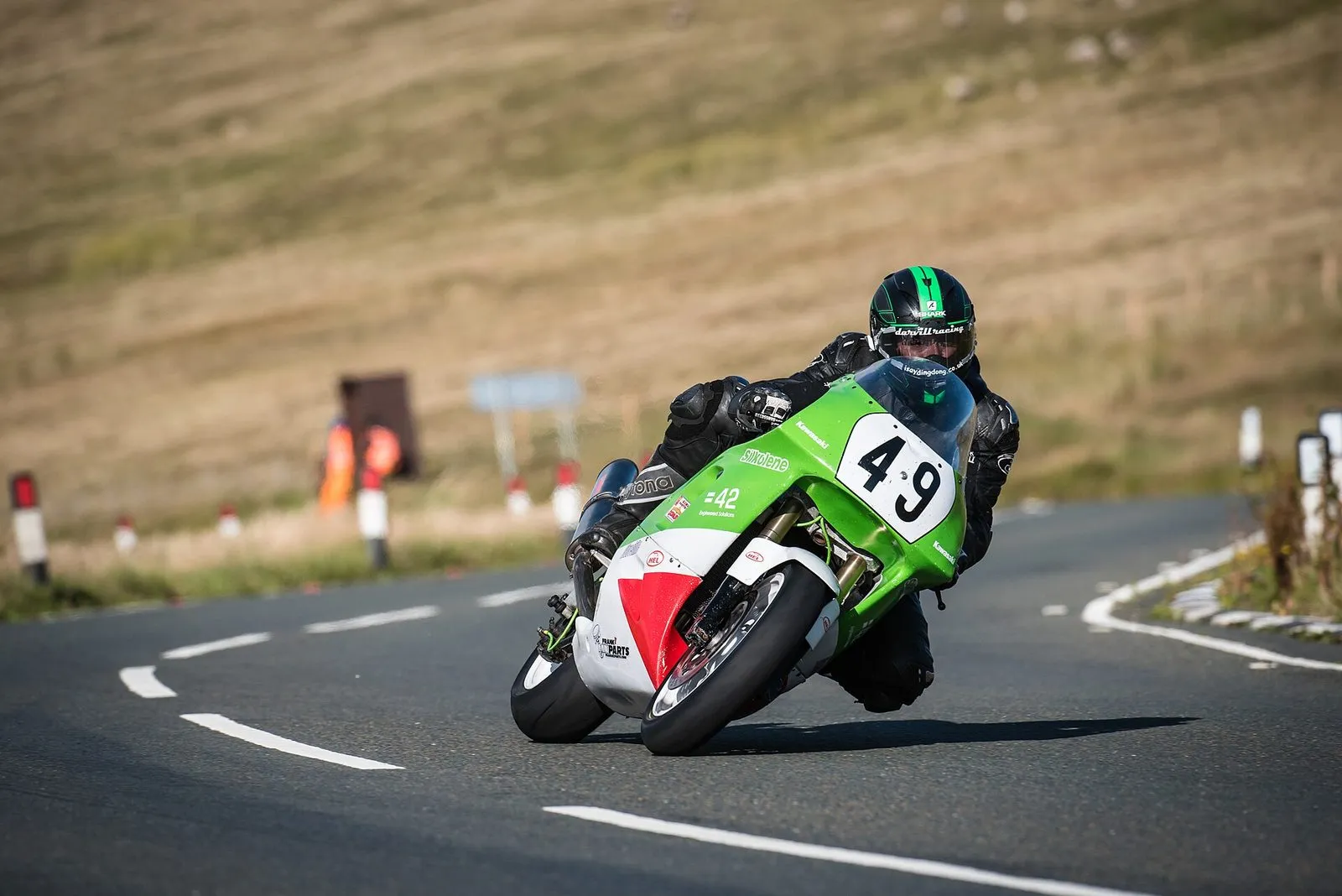 A Classic TT racer on a green and white bike leaning into the bend at Brandywell, with marshals and open moorland behind