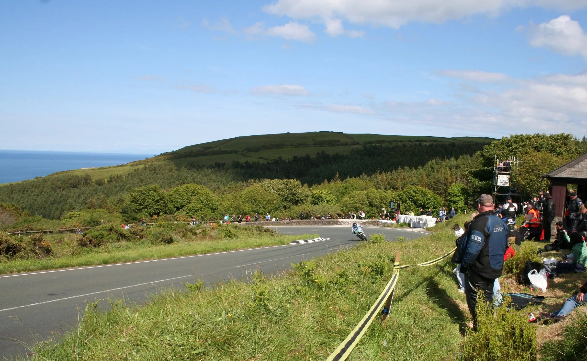 A racer leaning into the right-hand Gooseneck hairpin on the Mountain Road, watched by spectators on the bank, with North Barrule and the sea behind