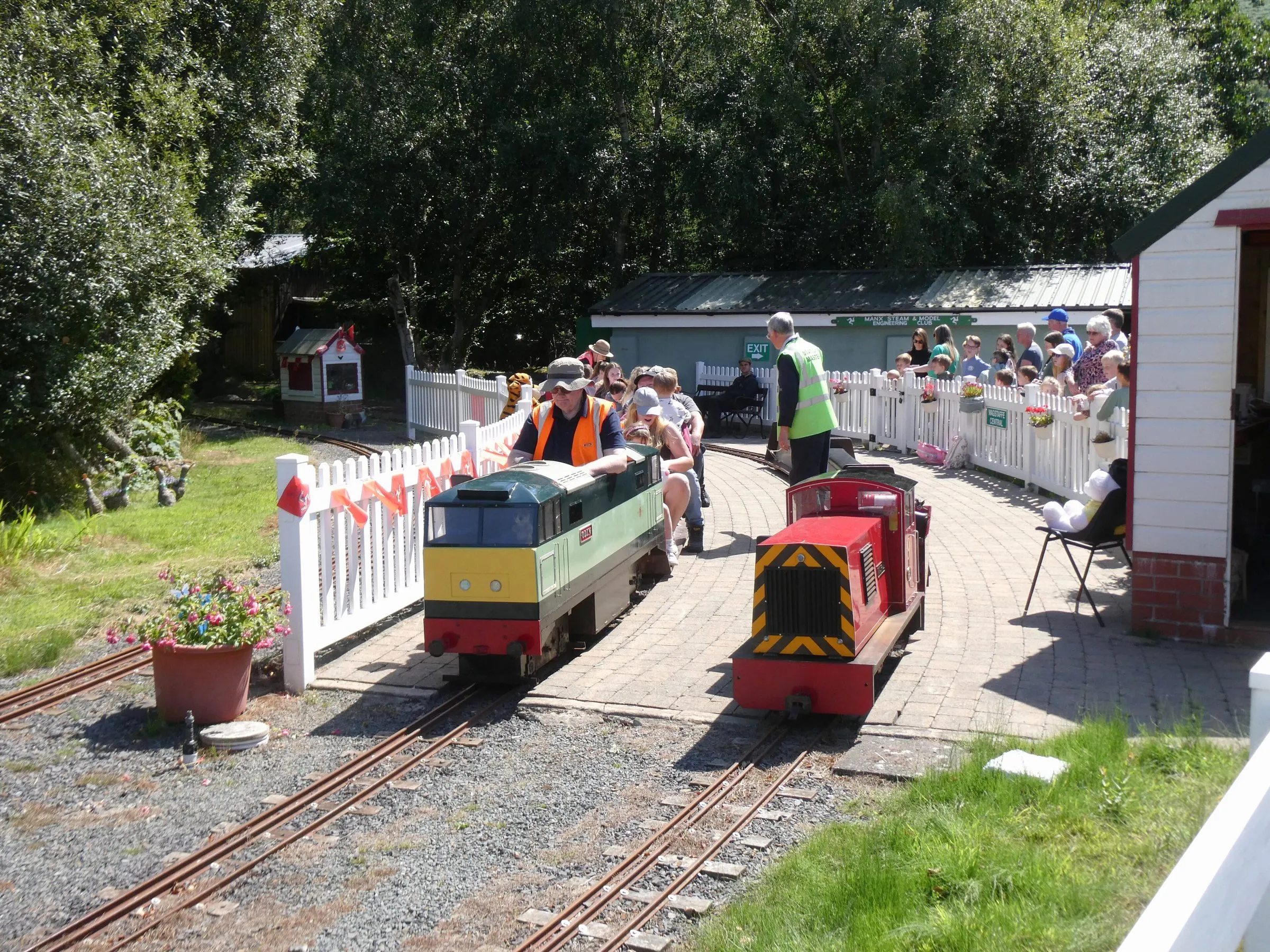 Two miniature locomotives at the Orchid Line station in Curraghs Wildlife Park, with families watching from behind a white picket fence on a sunny day
