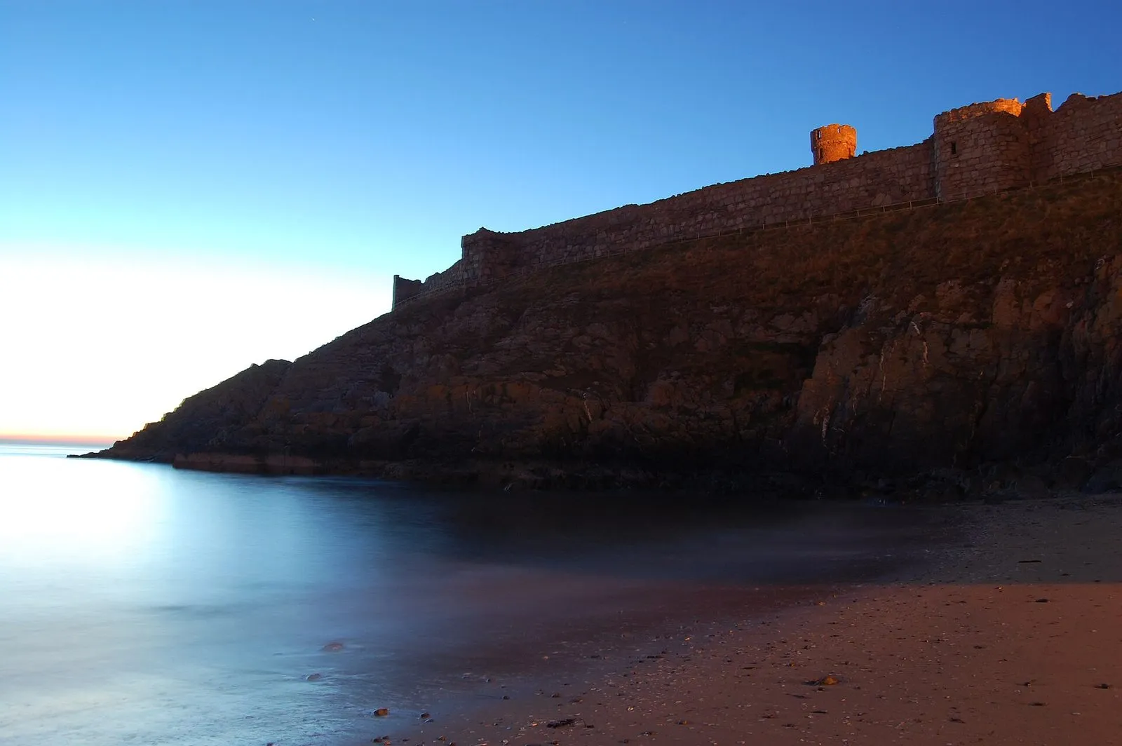 Peel Castle’s walls on St Patrick’s Isle lit by the last light, above a calm, blue-tinged sea at dusk