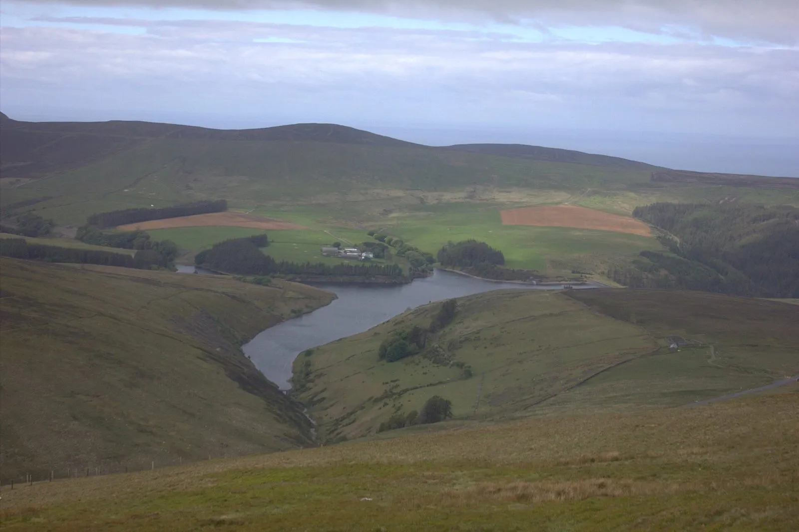 Sulby Reservoir winding through a green valley in the Manx hills, with Snaefell’s slopes around it and the sea on the horizon