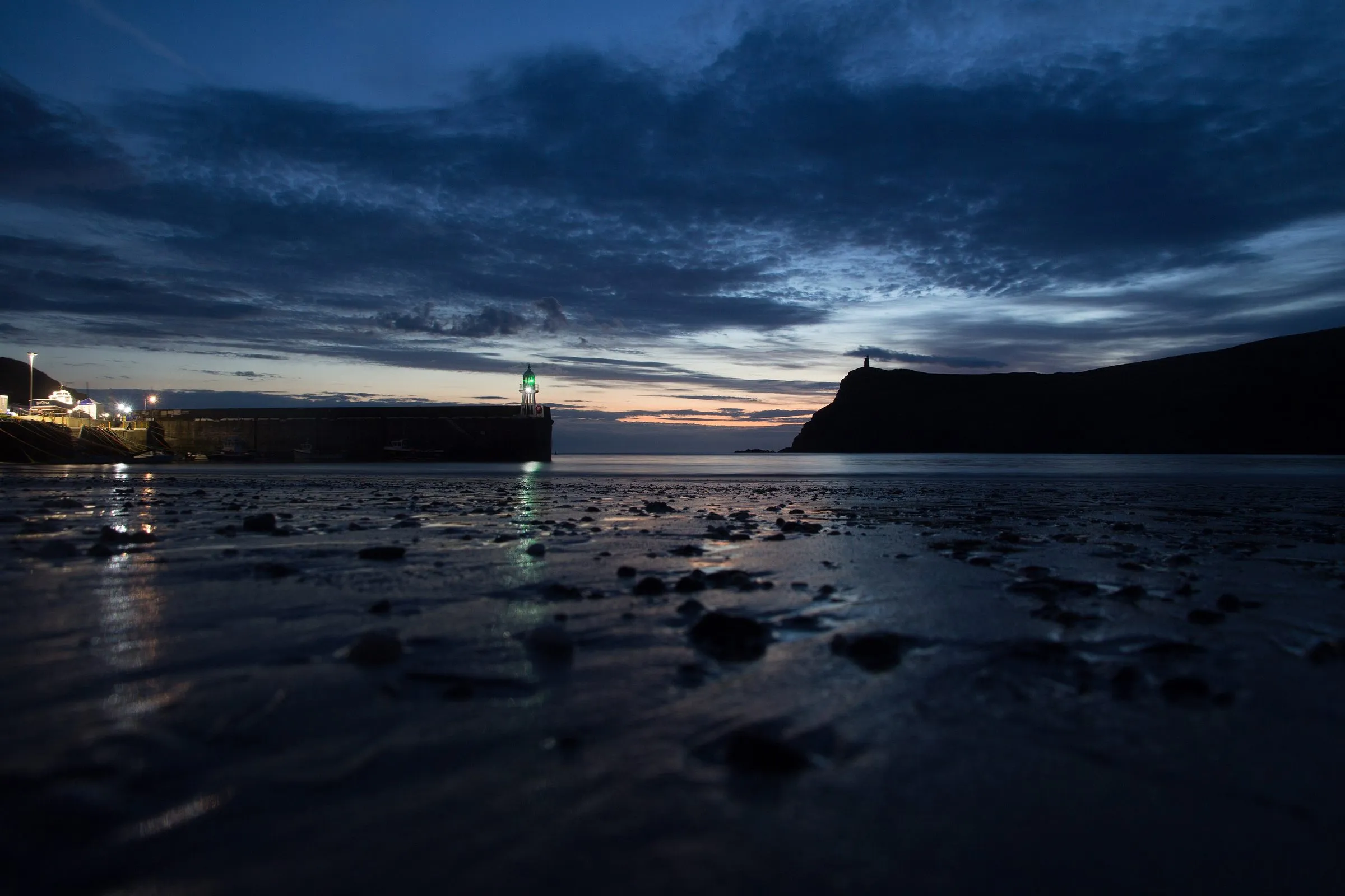 Port Erin bay at nightfall, the wet sand reflecting the harbour light, with Bradda Head and Milner's Tower in silhouette against a deep blue sky