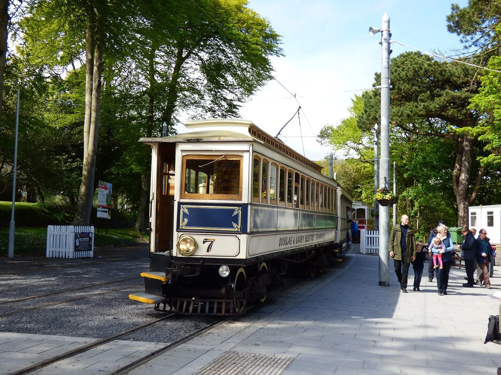 A cream and blue Manx Electric Railway tram, car number 7, at Laxey station under tall trees, with passengers on the platform