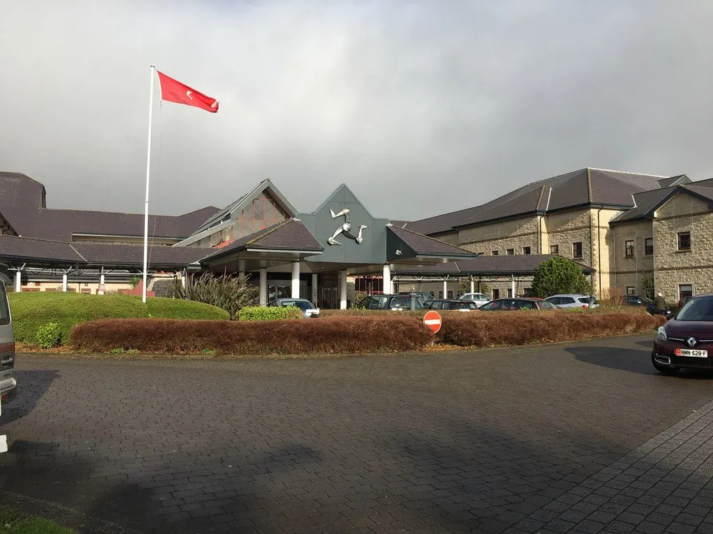 The main entrance of Noble’s Hospital, a low stone building with slate roofs and a three-legs sculpture over the door, with a Manx flag flying outside