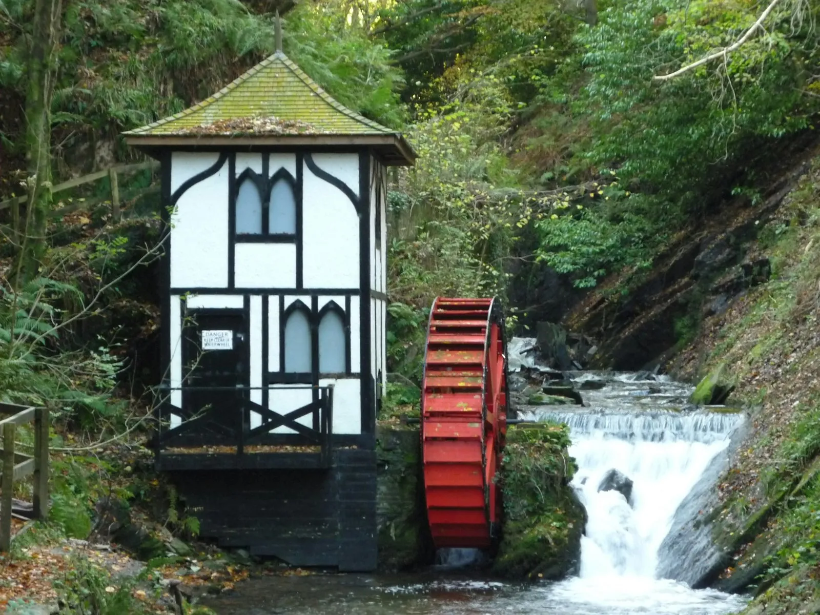 A small red waterwheel beside a black-and-white timbered hut and a waterfall in a wooded glen