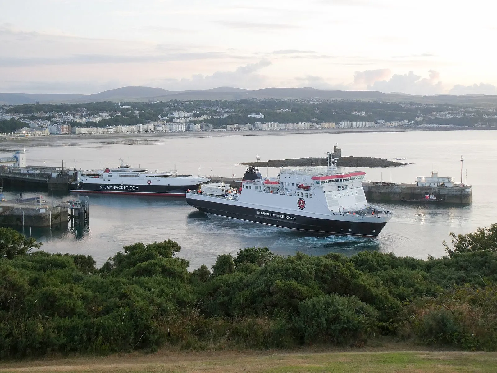 The conventional ferry Ben-my-Chree turning in Douglas Harbour beside the fast craft Manannan at her berth, with Douglas Bay and the hills behind