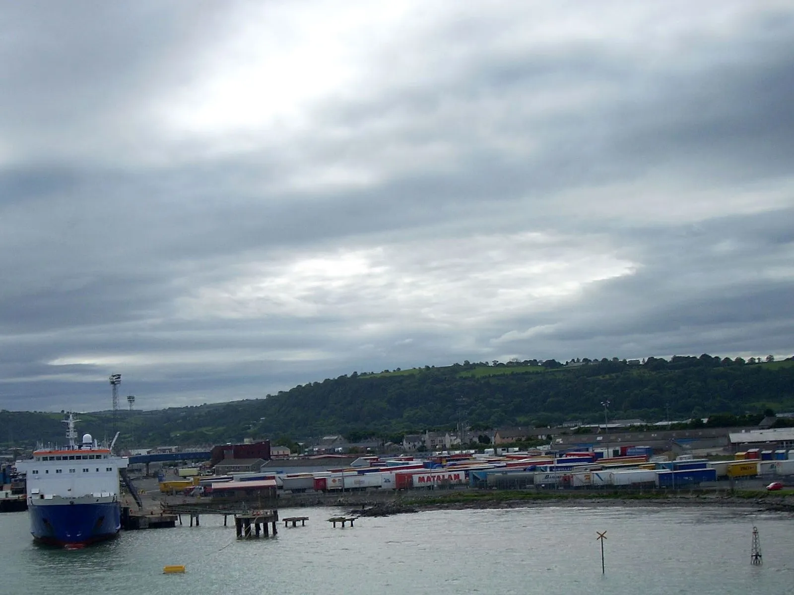 Larne harbour under heavy grey cloud, with a blue-hulled ferry at the berth on the left, lorry trailers lined up on the quay and wooded hills behind the town