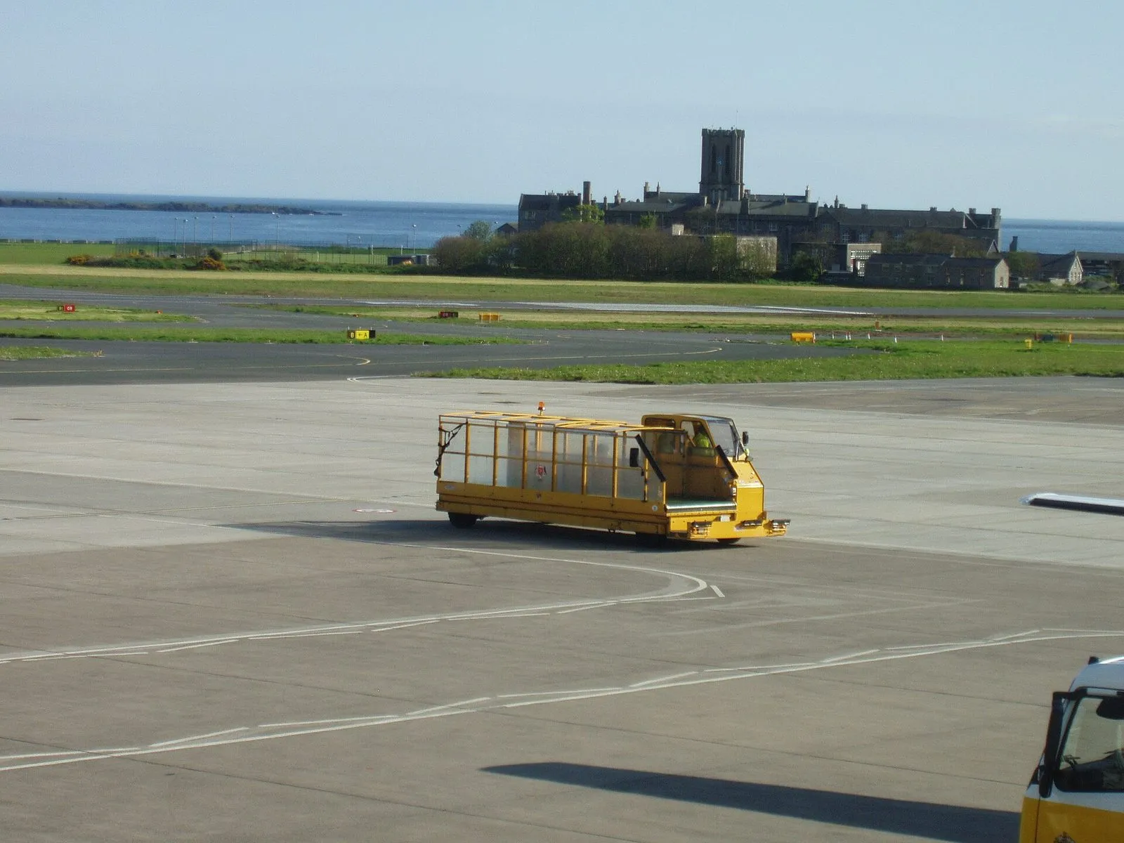A yellow baggage vehicle crossing the apron at Ronaldsway, with the tower of King William’s College and the sea behind