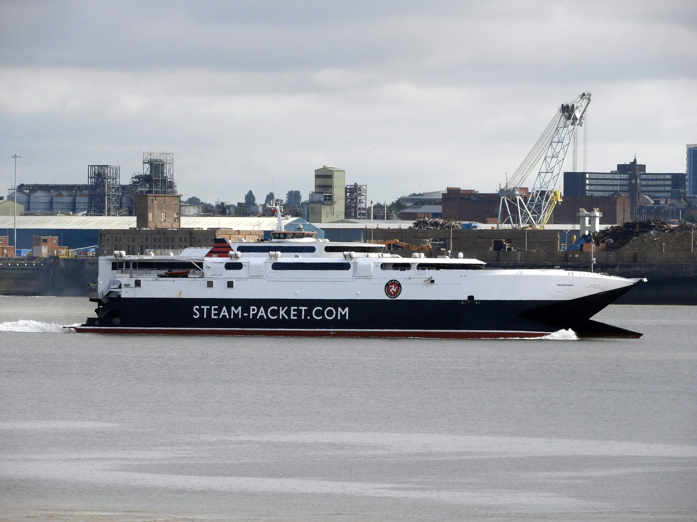 The Steam Packet fast craft Manannan, white with a dark hull marked steam-packet.com, speeding up the Mersey past cranes and warehouses