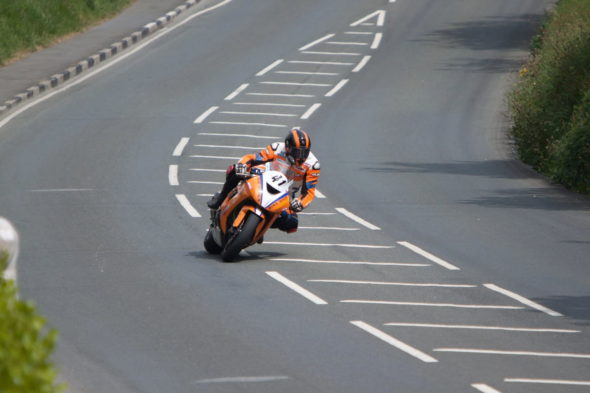 A rider in orange leathers leaning a sports bike through Bedstead Corner on the TT course, a Manx road between hedges