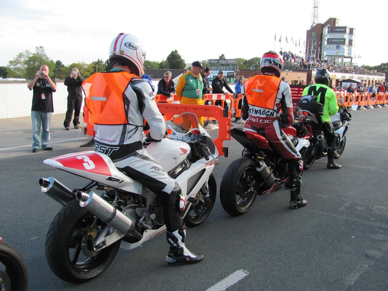 Manx Grand Prix riders in orange bibs lined up on their bikes at the Grandstand before a session, with marshals and spectators behind the barriers