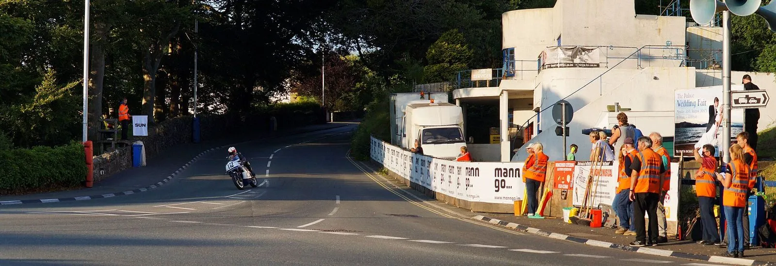 Marshals in orange tabards behind a barrier at a corner as a rider comes through during Manx Grand Prix practice on a summer evening