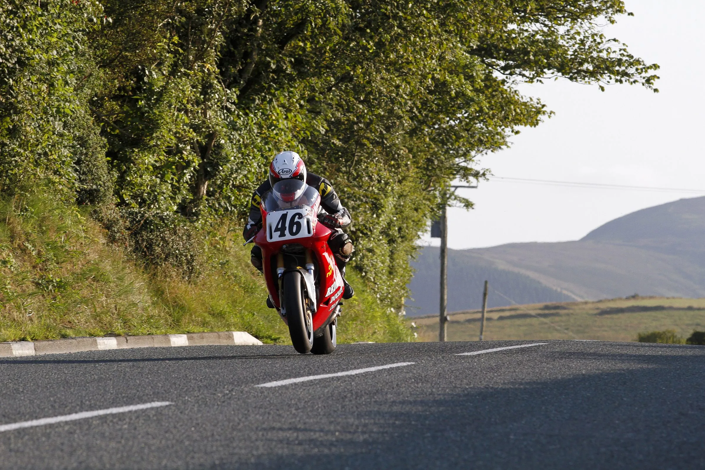 A rider on a red Ducati, number 46, leaning out of a tree-lined bend at Lambfell on the TT course, with bare hills behind