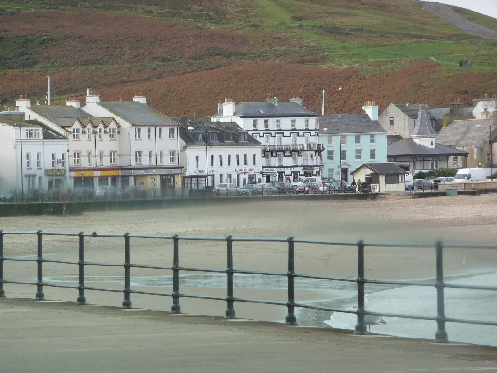 The seafront at Peel, with a row of white and pale-blue buildings along the promenade behind the empty beach and a heather-covered hill above