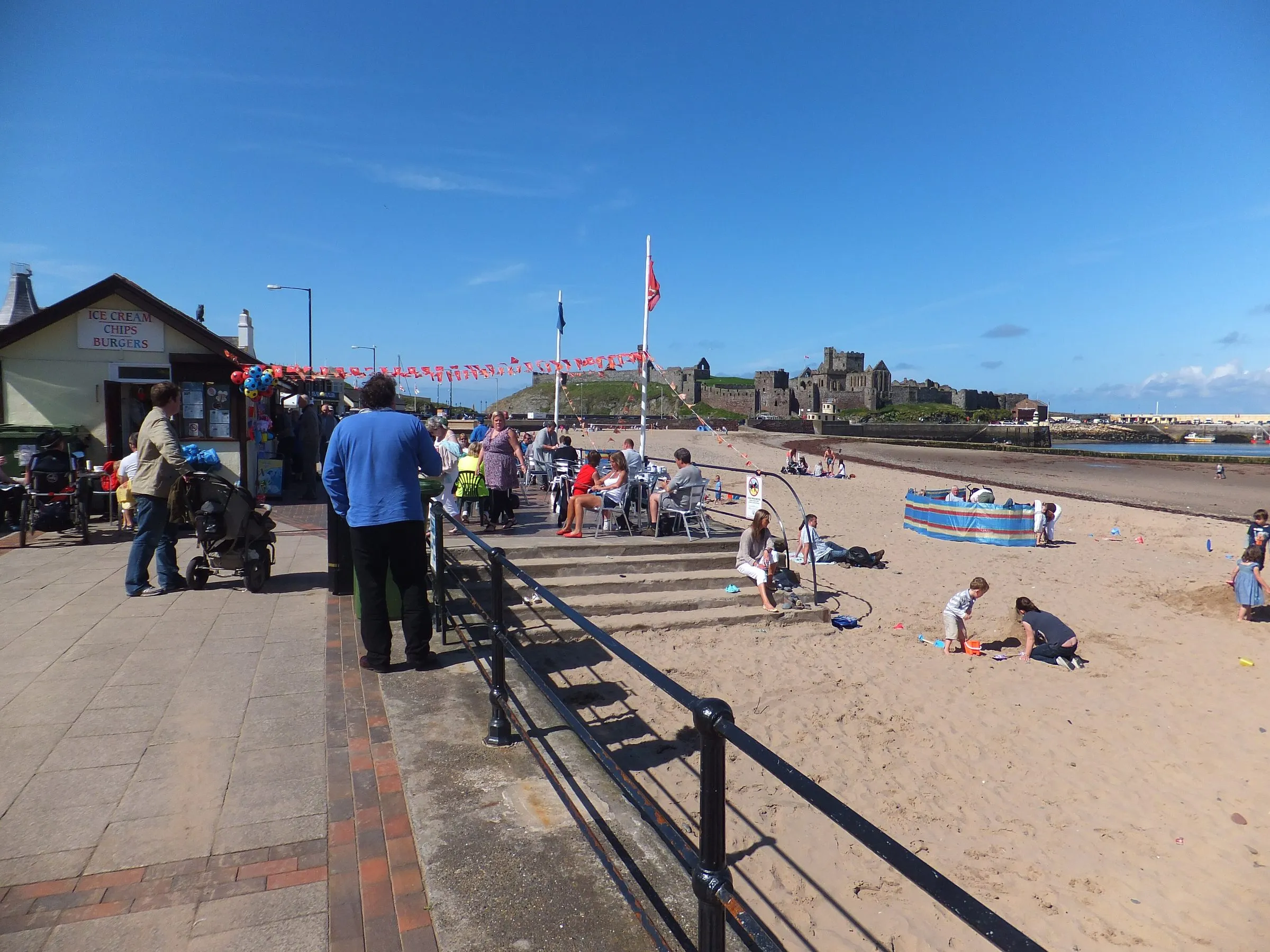 People on the sunny promenade at Peel beside a kiosk advertising ice cream, chips and burgers, with the beach and Peel Castle beyond