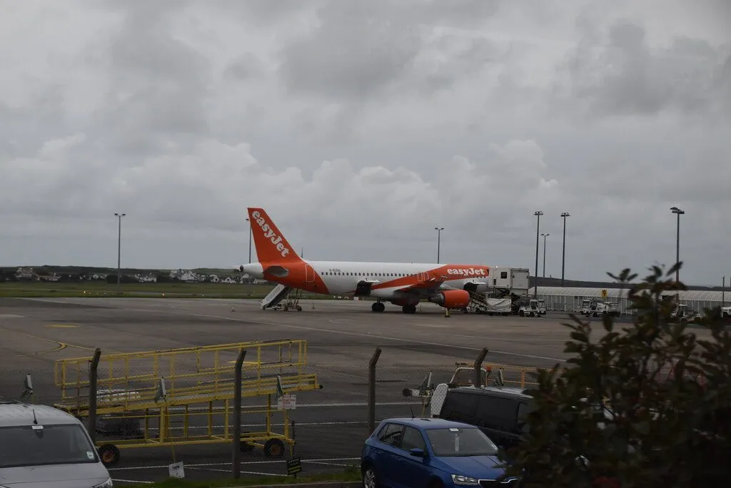 An easyJet Airbus parked on the apron at Ronaldsway under a grey sky, with the green fields of the south of the island behind