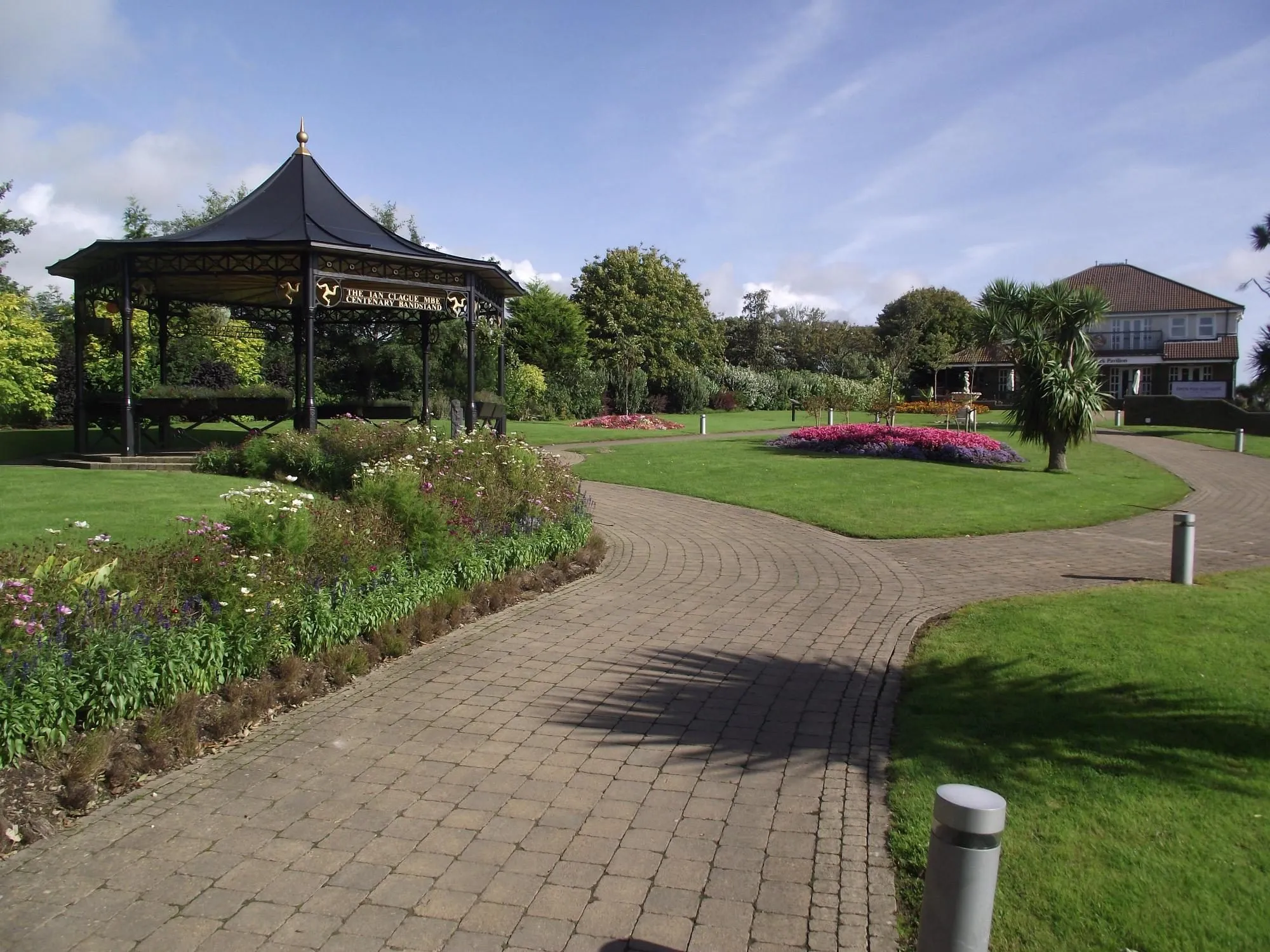 A black bandstand and flowerbeds beside a curving brick path in Noble's Park, Douglas, with the park pavilion behind under a blue sky