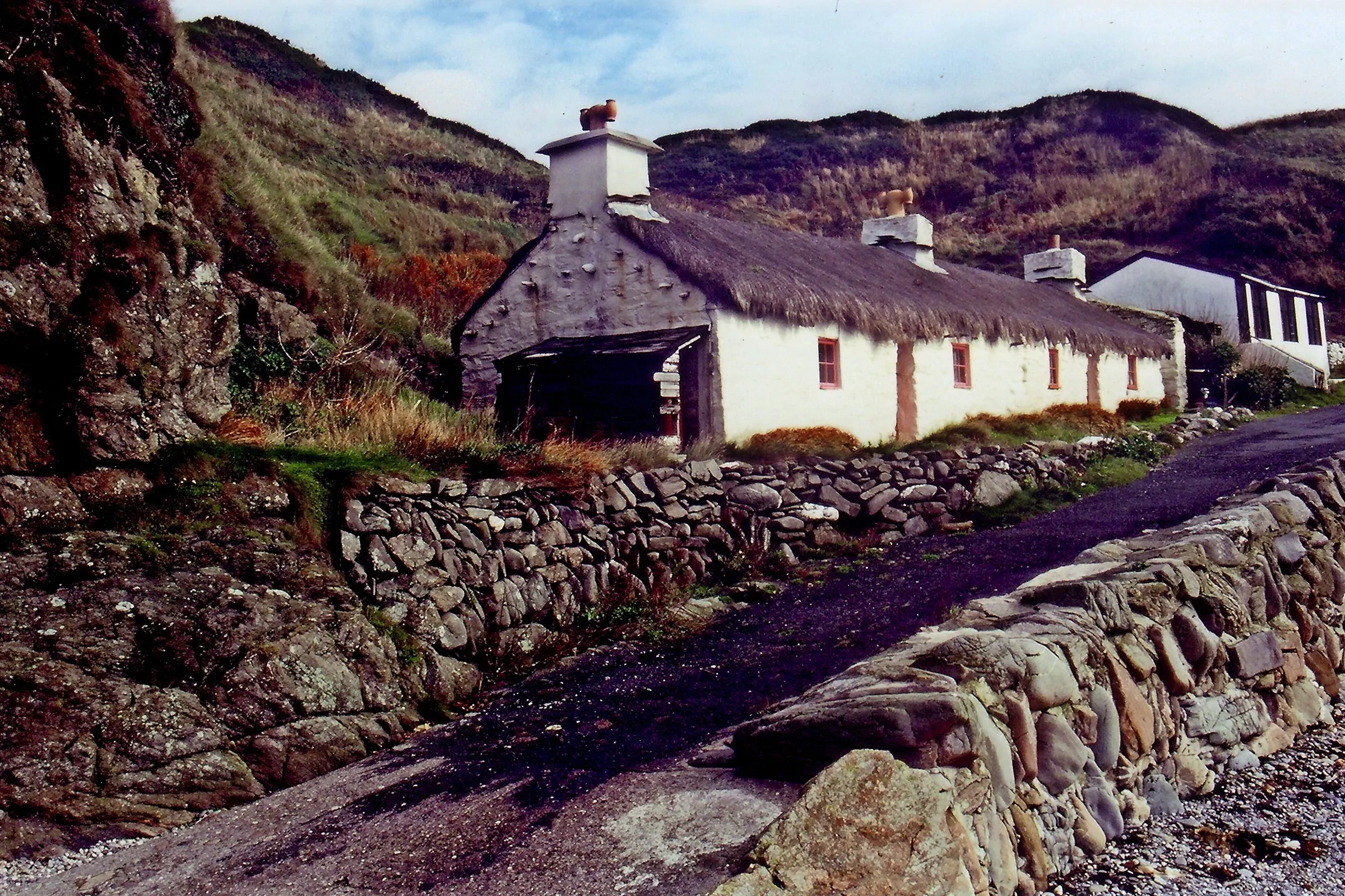 The whitewashed thatched fisherman's cottage at Niarbyl beside a stone wall and the shore, with bracken-covered hills behind
