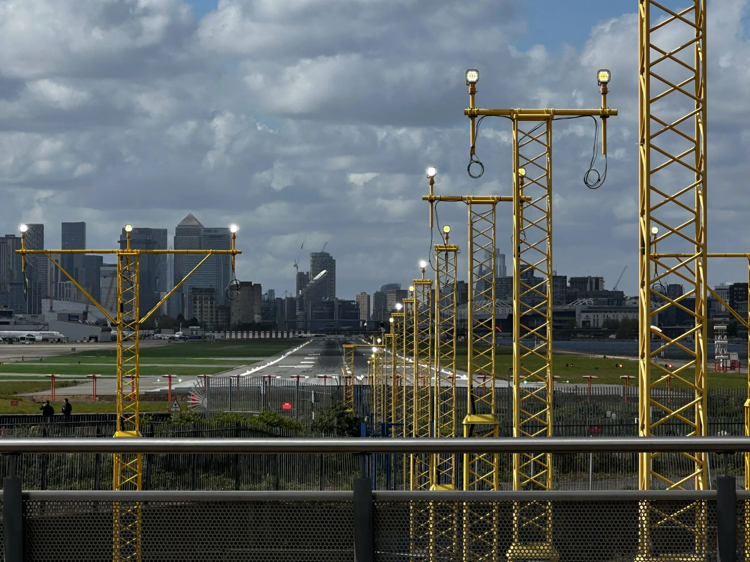 The runway at London City Airport lined with yellow approach-light towers, with the Canary Wharf skyline behind under a cloudy sky