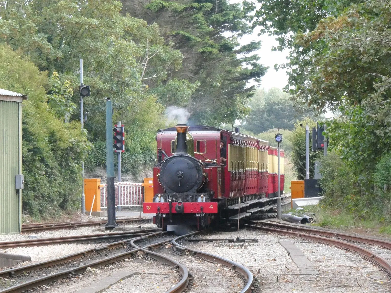 A red steam locomotive pulling a rake of red and cream carriages into Port Erin station between trees