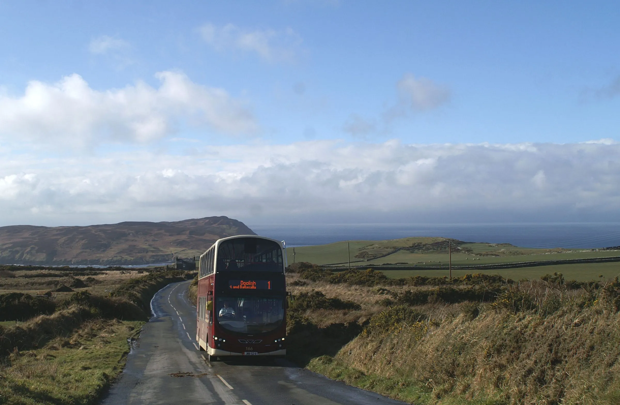 A red double-decker Bus Vannin number 1 to Douglas on a narrow lane near Cregneash, with the sea and the Calf of Man behind