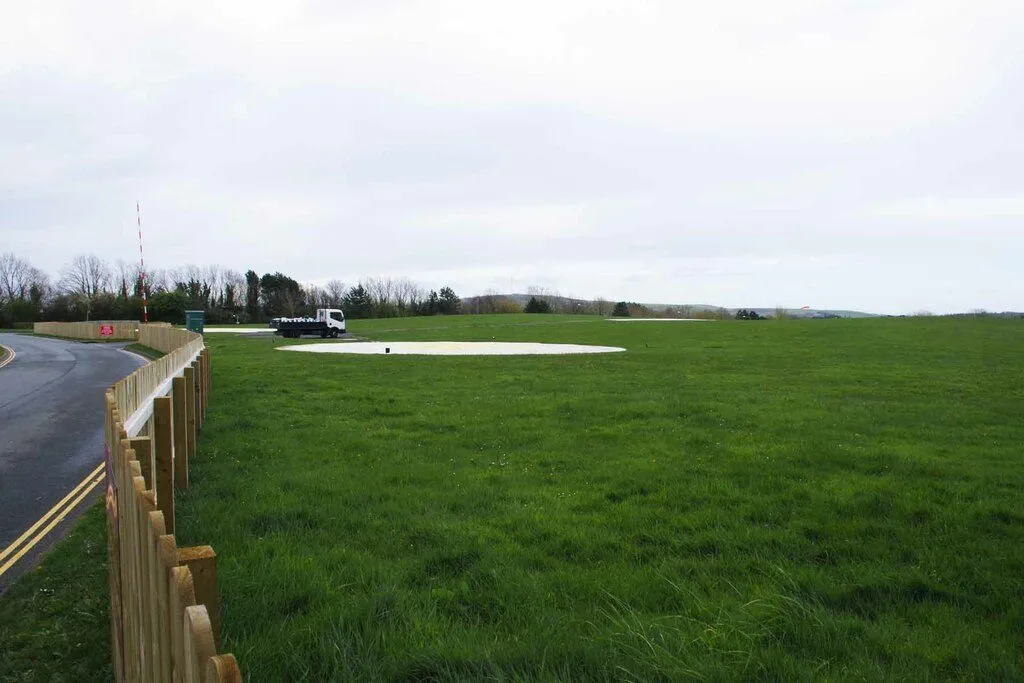 Two white helicopter landing pads on a wide grass field beside the road into Noble’s Hospital, under a grey sky