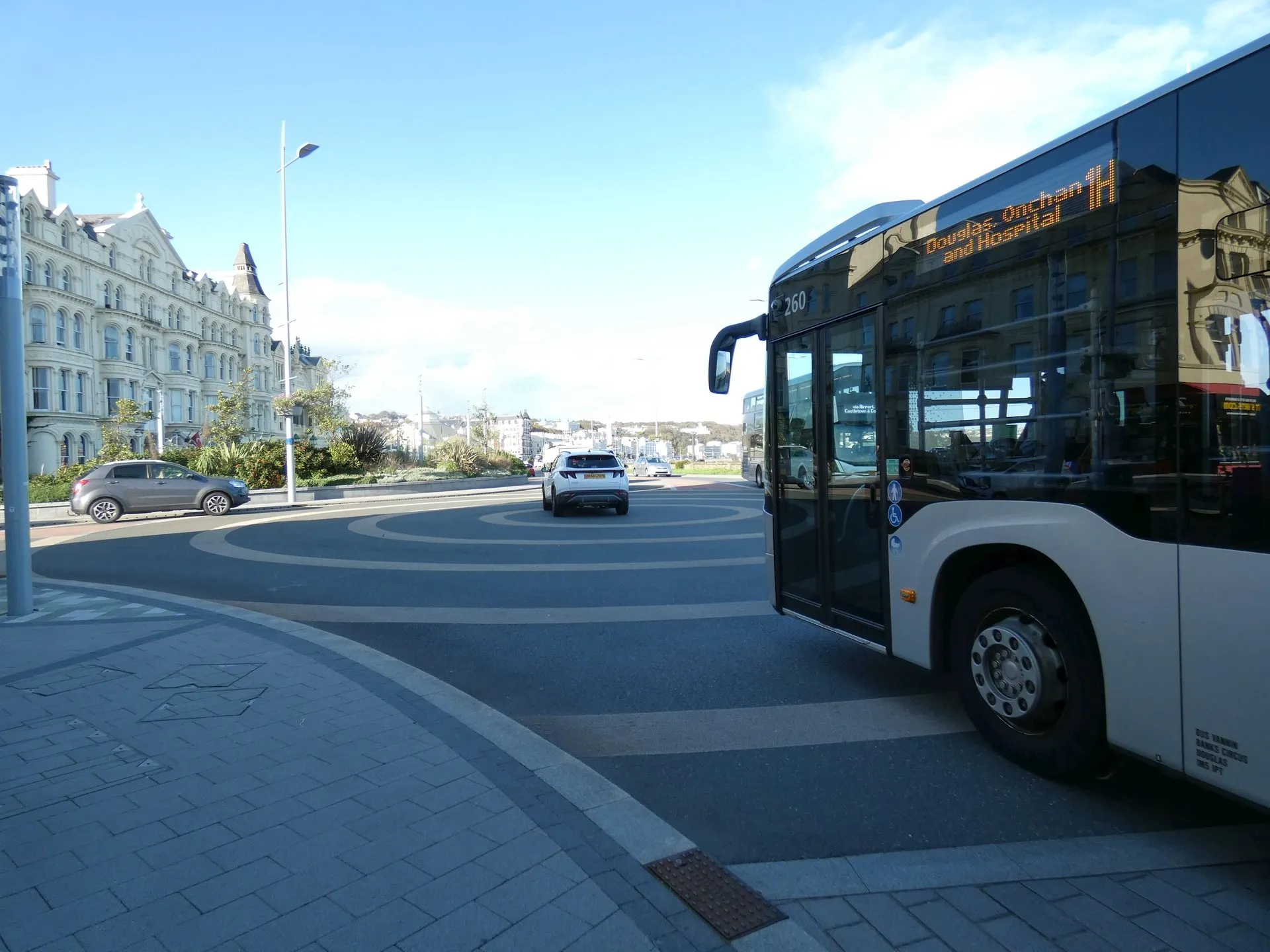 A Bus Vannin bus showing route 1H to Douglas, Onchan and Hospital, pulling away along Douglas promenade under a blue sky