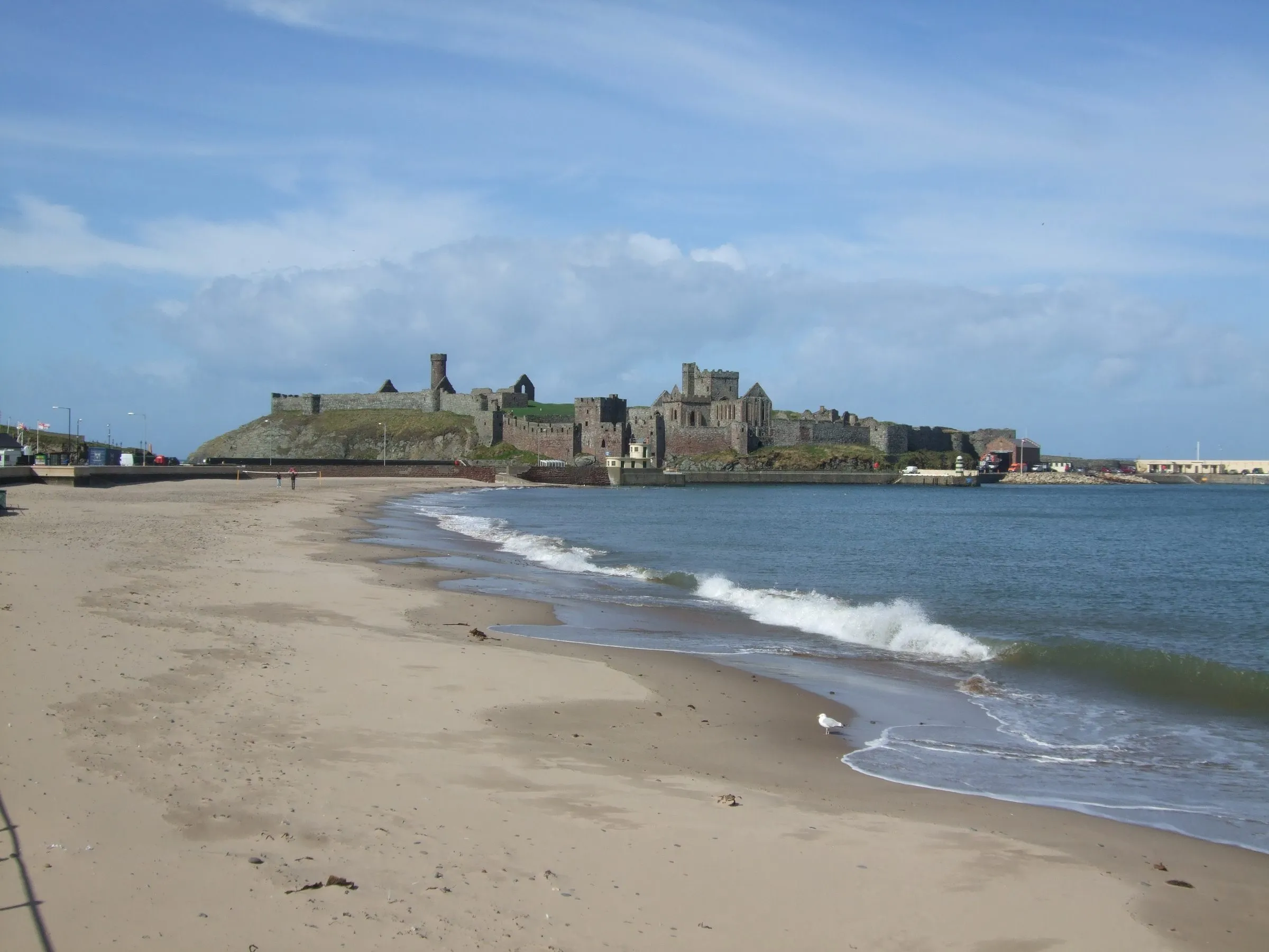 Peel Castle's red sandstone walls on St Patrick's Isle, seen along the curve of Peel beach with waves breaking on the sand