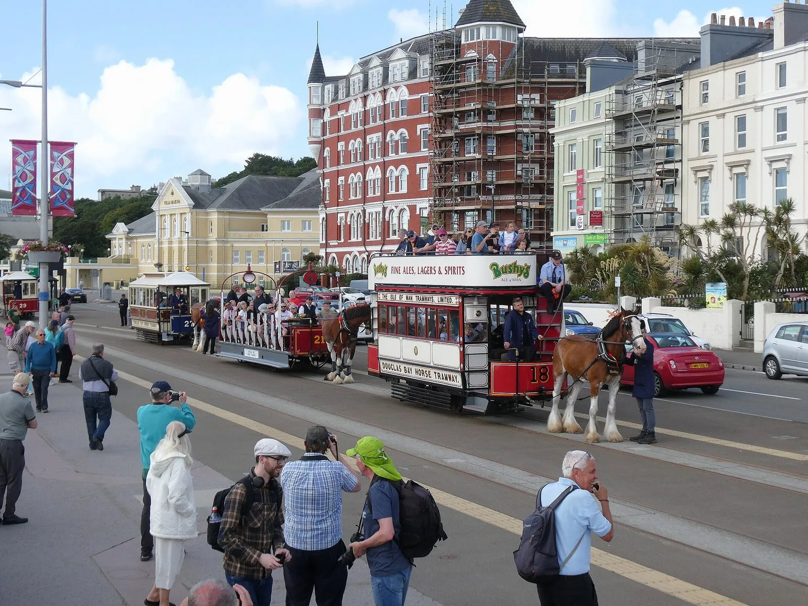 Horse trams in a cavalcade on Douglas promenade: a double-deck tram numbered 18 pulled by a horse, with open trams behind and a crowd watching from the pavement