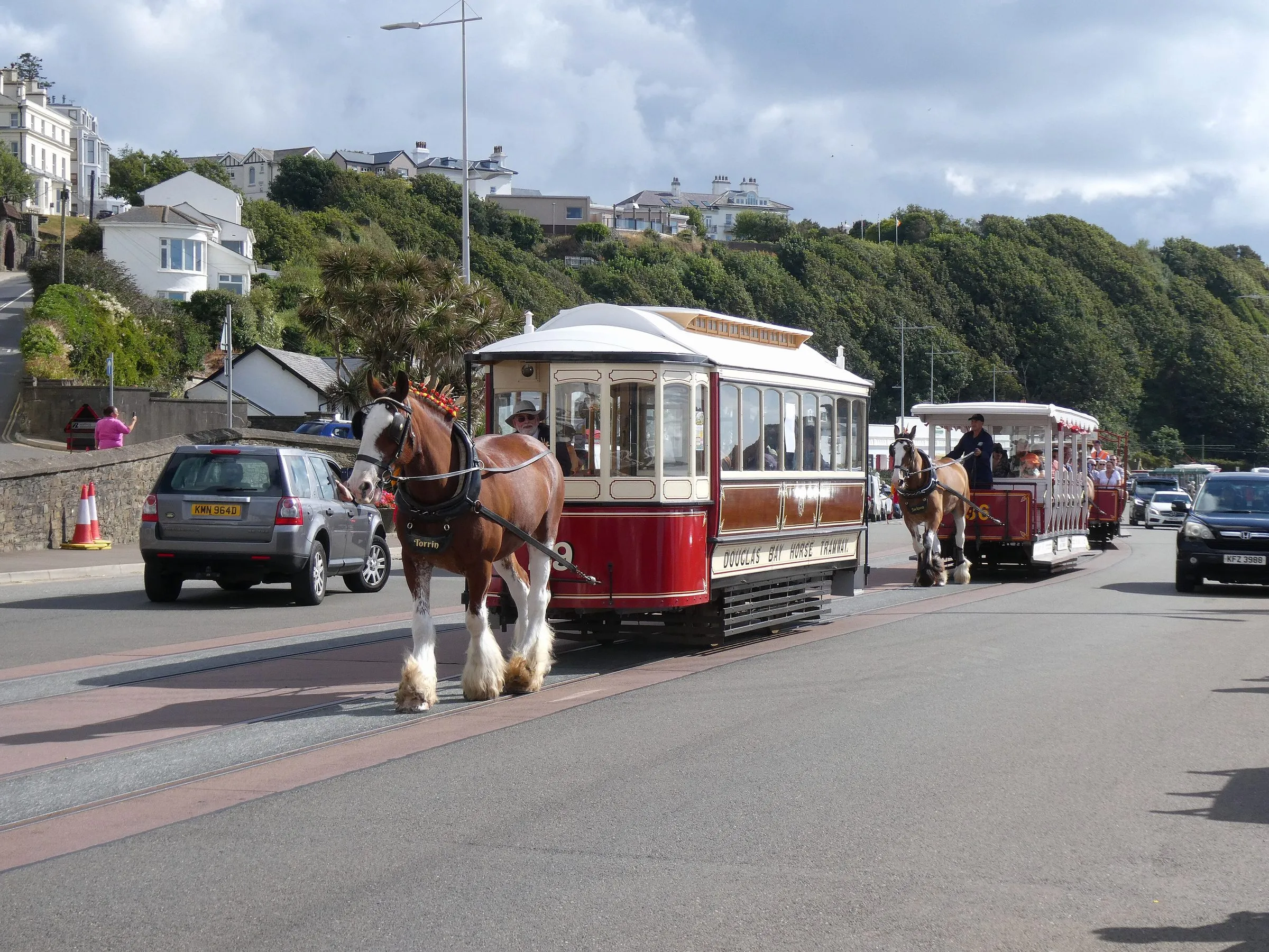 A Clydesdale horse pulling a red and cream closed horse tram along the centre of Douglas promenade, with an open toastrack tram following behind